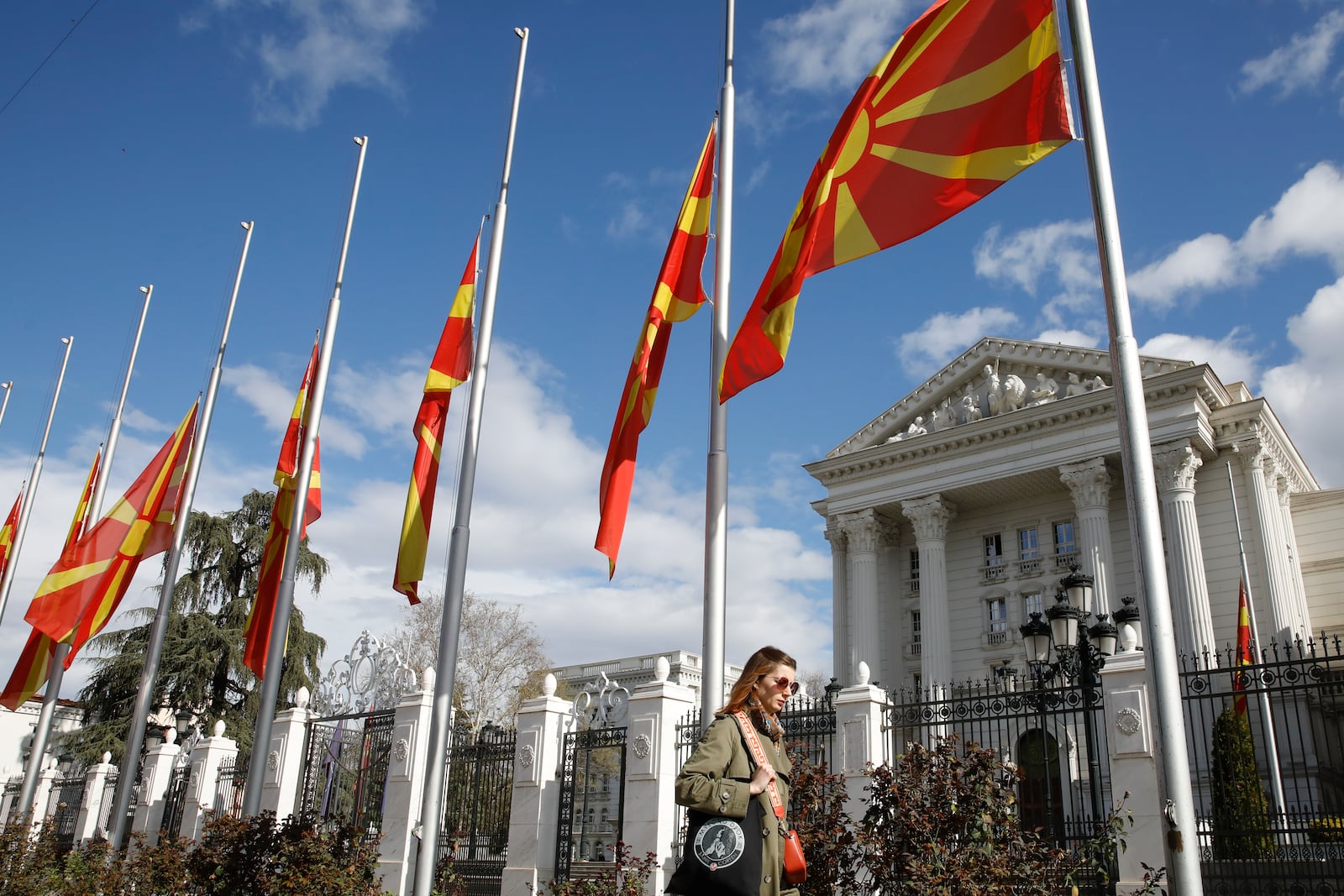 A woman walks past North Macedonia flags flapping in the wind at half staff in front of a Government building in Skopje, North Macedonia, Monday, March 17, 2025, following a massive fire in the nightclub in the town of Kocani, early Sunday. (AP Photo/Boris Grdanoski)