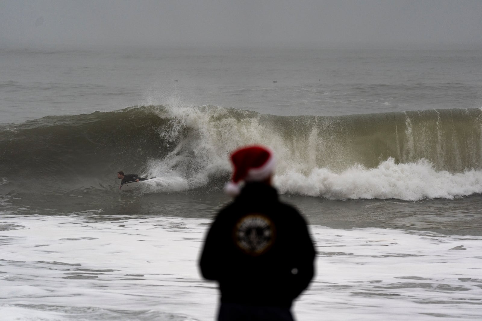 Jeff Parker, wearing a Santa Claus hat, watches as a surfer rides a wave in Seal Beach, Calif., Tuesday, Dec. 24, 2024. (AP Photo/Jae C. Hong)