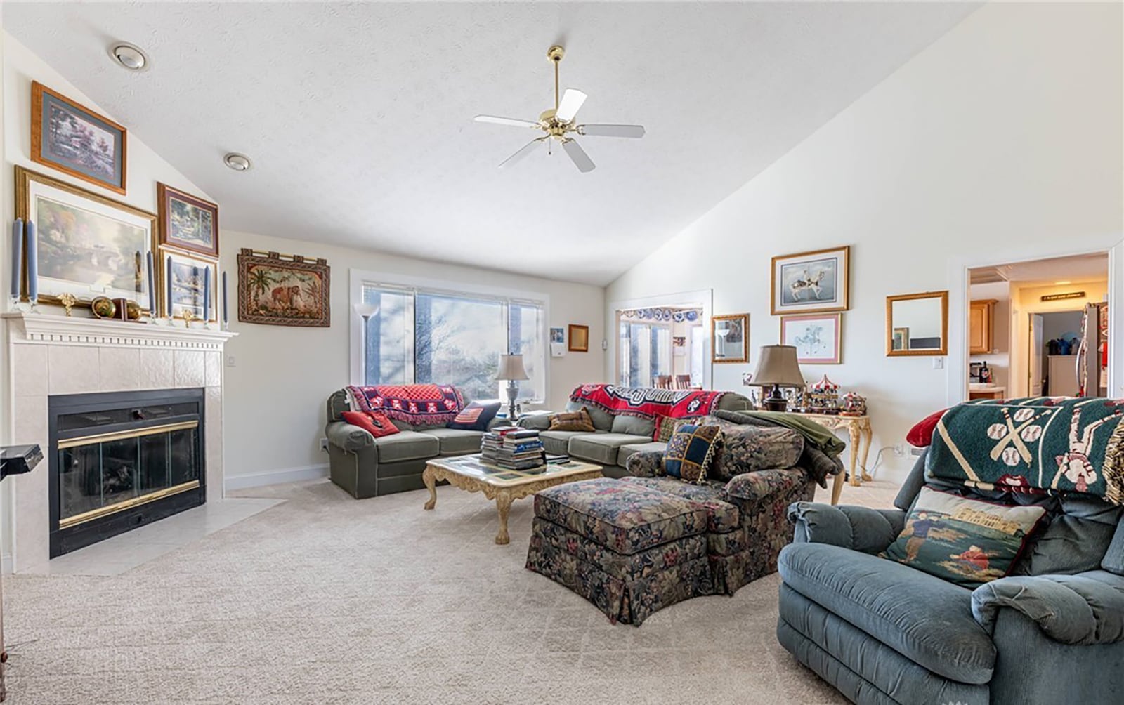 The great room has a gas fireplace, vaulted ceiling, neutral carpeting and a ceiling fan.