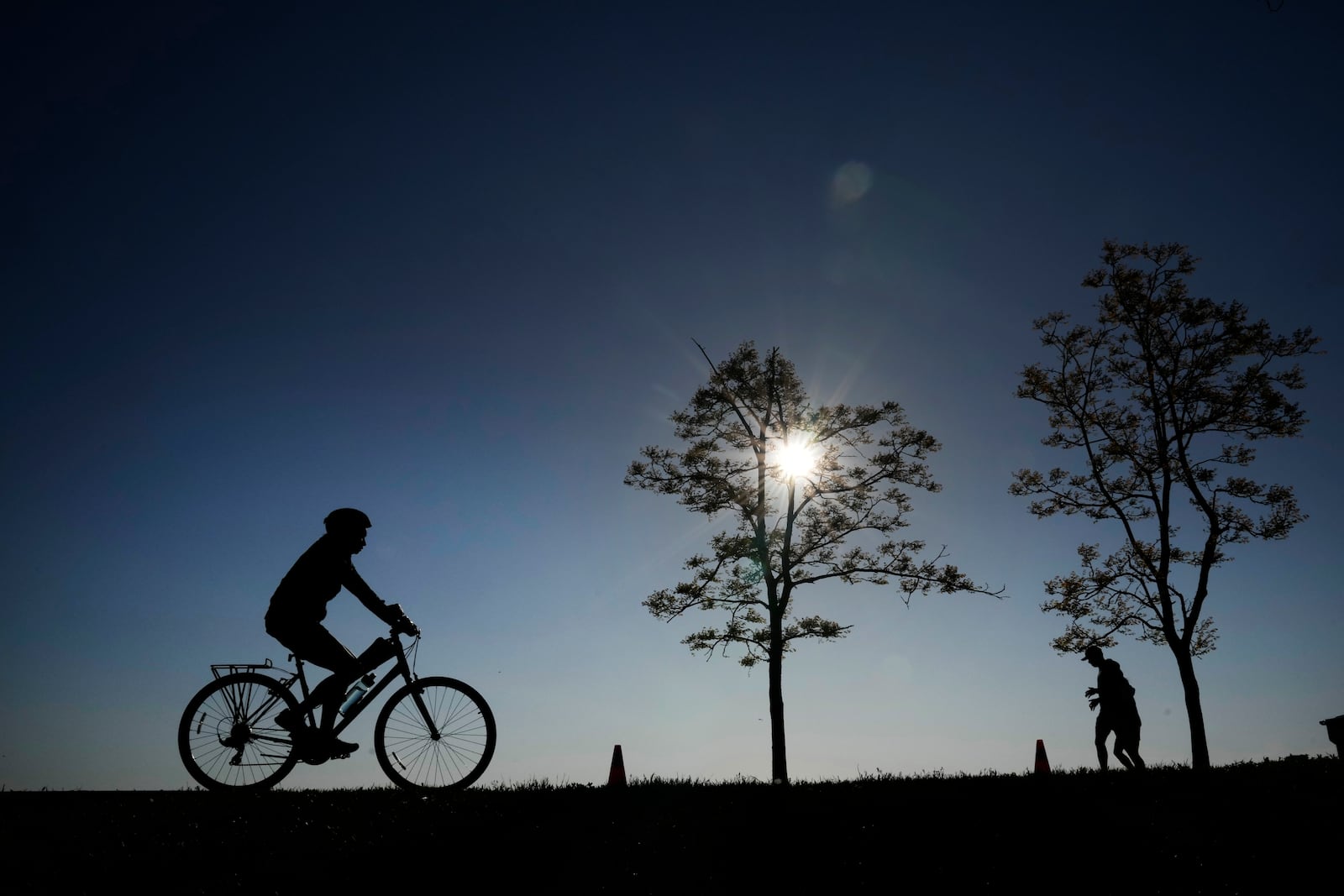 FILE - A cyclist and two joggers in silhouette navigate the bike path along Lake Michigan at the 31st Street Harbor Saturday, May 27, 2023, in Chicago. (AP Photo/Charles Rex Arbogast, File)