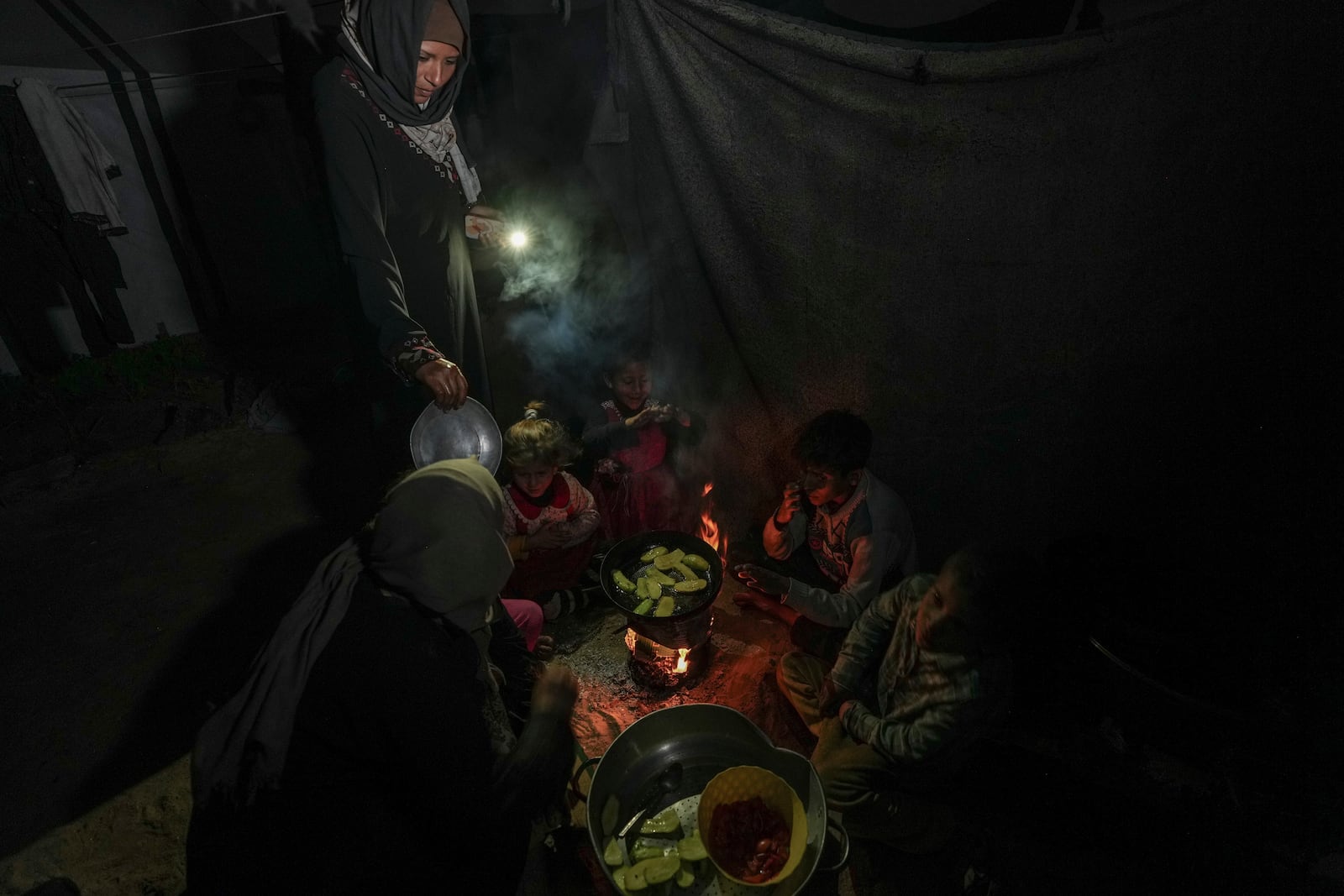 Reda Abu Zarada, left, and her daughter, Amani, standing, displaced from Jabaliya in northern Gaza, feed their children and grandchildren with fried zucchini made over a fire made of paper and cardboard scraps outside their tent in a camp in Khan Younis, Gaza Strip, Thursday, Dec. 19, 2024. (AP Photo/Abdel Kareem Hana)