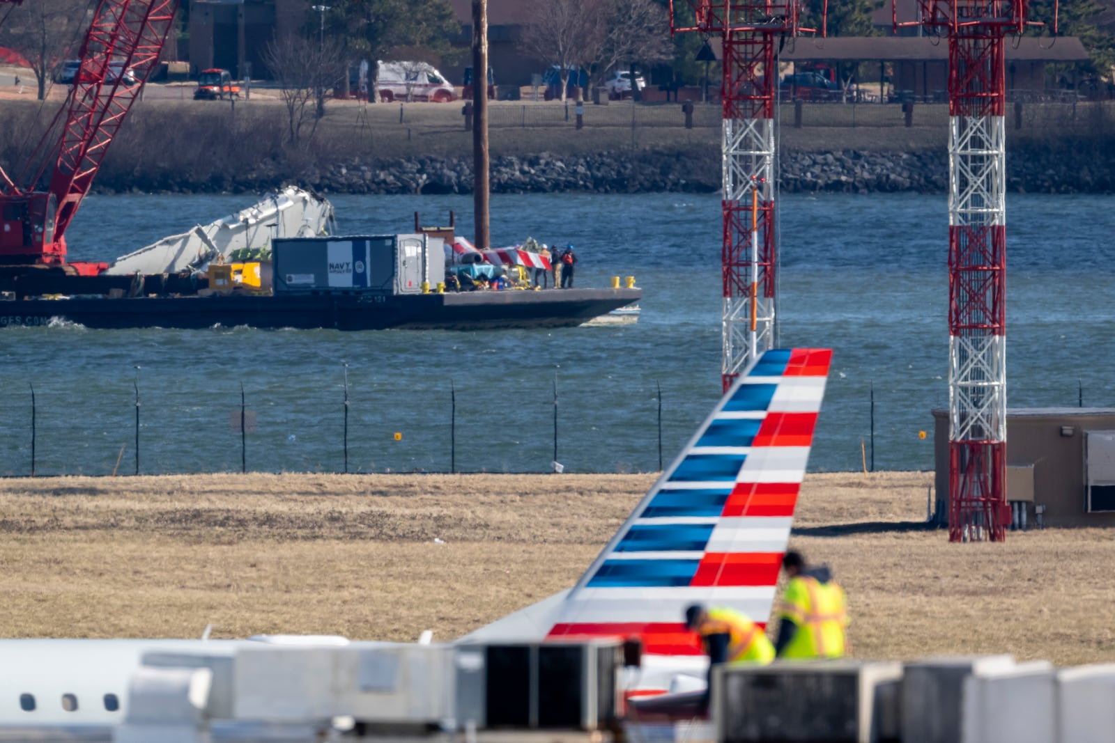 Parts of a plane are lifted from the water near the wreckage site in the Potomac River of a mid-air collision between an American Airlines jet and a Black Hawk helicopter, at Ronald Reagan Washington National Airport, Tuesday, Feb. 4, 2025, in Arlington, Va., as an American Airlines aircraft is seen landing on the runway, foreground. (AP Photo/Ben Curtis)