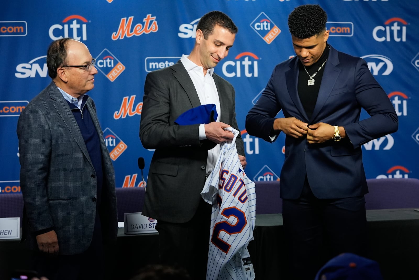 New York Mets owner Steven Cohen, left, looks on as new Mets plyaer Juan Soto, right, is presented with a jersey by the team's president of baseball operations David Stearns, center, during a news conference, Thursday, Dec. 12, 2024, in New York. (AP Photo/Frank Franklin II)