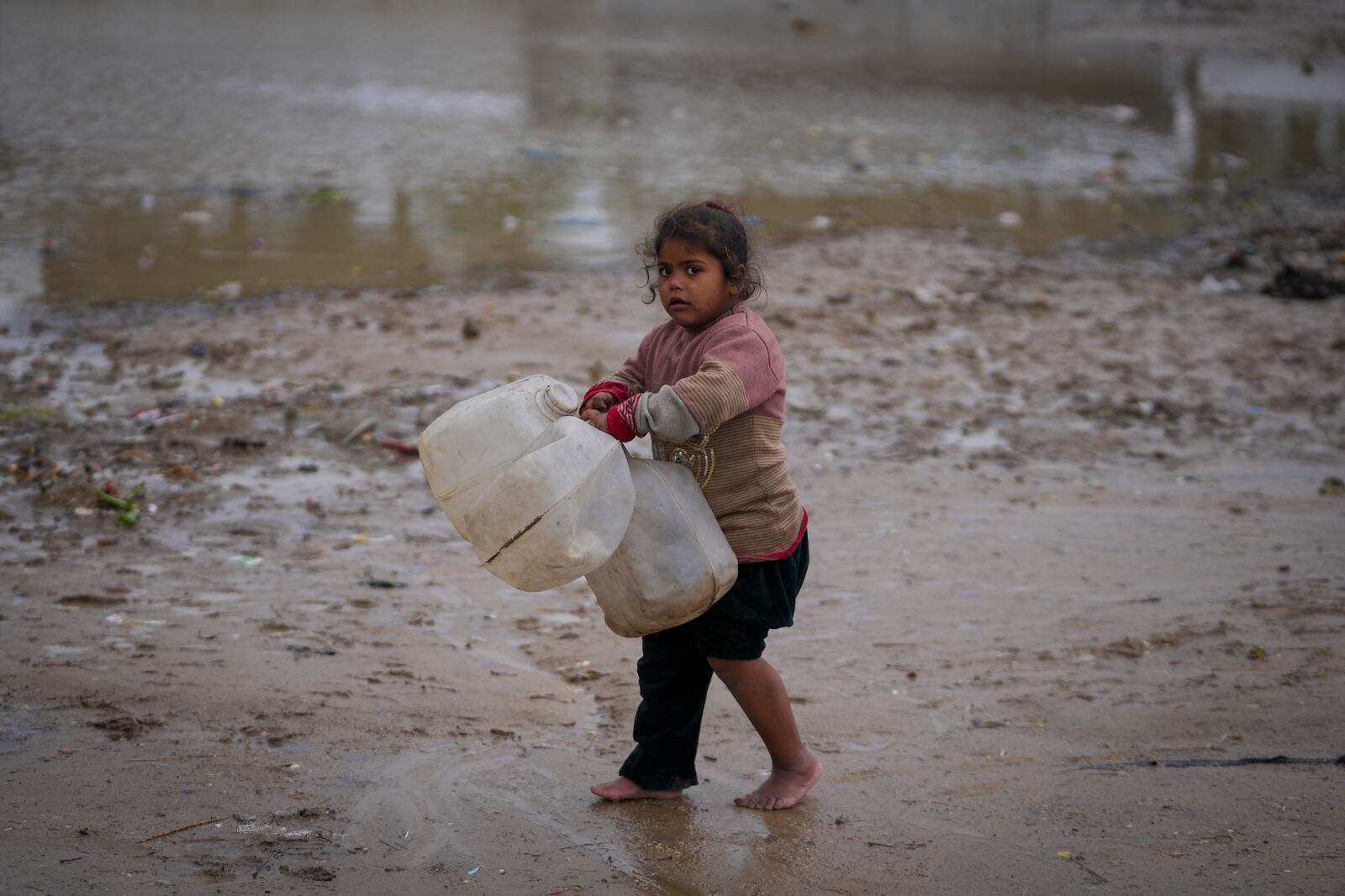 A young girl walks barefoot, carrying empty jerrycans to collect water, after overnight rainfall at the refugee tent camp for displaced Palestinians in Deir al-Balah, central Gaza Strip,, Tuesday, Dec. 31, 2024. (AP Photo/Abdel Kareem Hana)