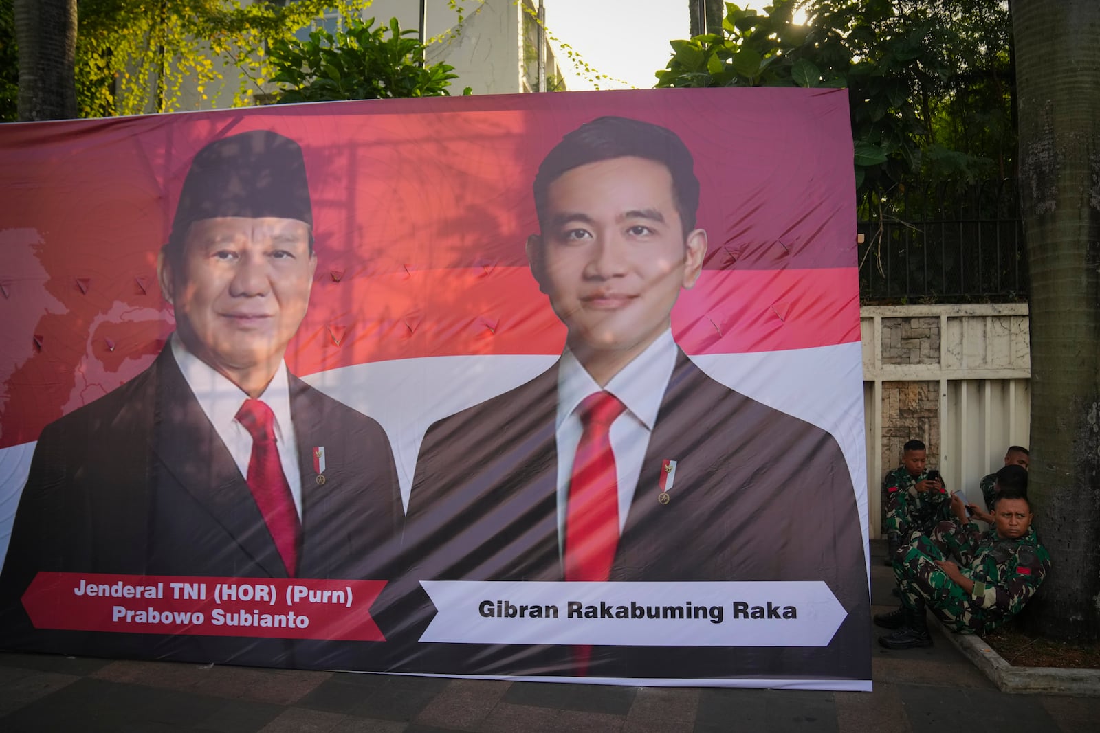 Soldiers sit next to a banner showing Indonesian President-elect Prabowo Subianto, left, and Vice President-elect Gibran Rakabuming Raka, who is also the eldest son of outgoing President Joko Widodo in Jakarta, Indonesia, Sunday, Oct. 20, 2024. (AP Photo/Dita Alangkara)