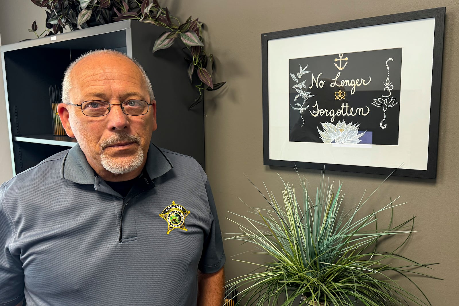 Hamilton County Coroner Jeff Jellison stands in his office in Noblesville, Indiana, July 11, 2024, in front of a painting reading "No Longer Forgotten" that his wife created as reminder of his ongoing work to identify some 10,000 human bones and bone fragments unearthed on Herbert Baumeister's suburban Indianapolis property starting in 1996. (AP Photo/Rick Callahan)