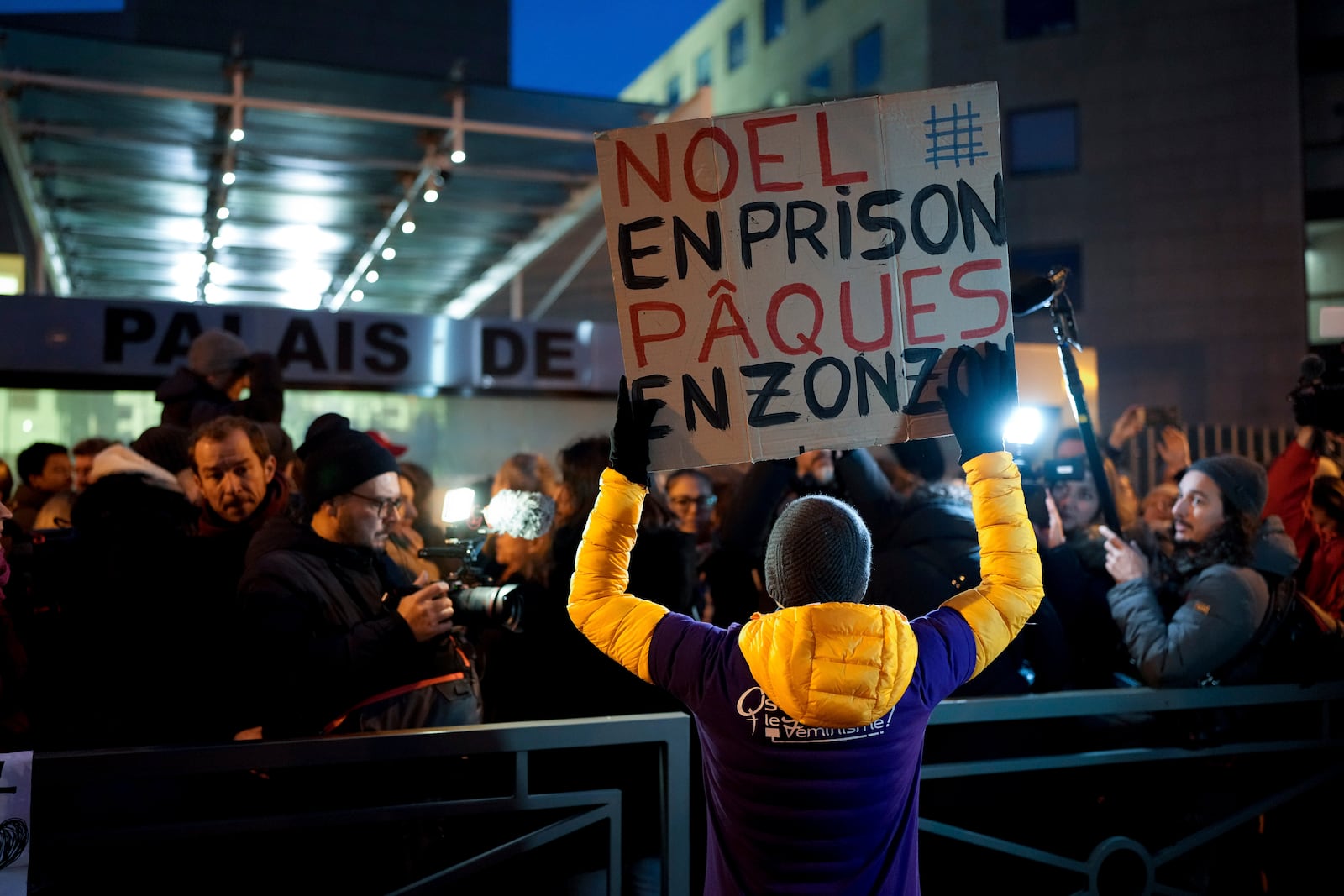 A man with a placard reading "Christmas in prison, Easter in the slammer" walks past the media as they wait outside the courthouse of Avignon during the trial of Dominique Pelicot, in Avignon, southern France, Thursday, Dec. 19, 2024. (AP Photo/Lewis Joly)