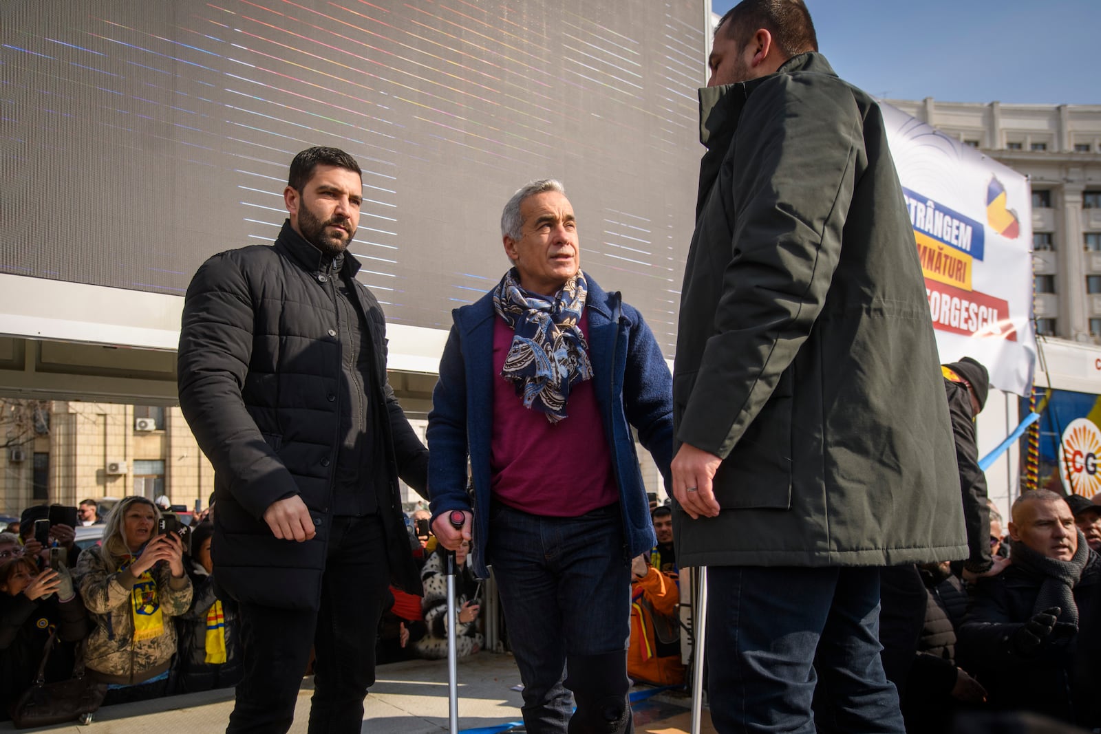 Calin Georgescu, the winner of Romania's first round of presidential election, annulled by the Constitutional Court, uses crutches while standing on a stage to greet supporters gathered for a protest outside the Romanian parliament in Bucharest, Romania, Saturday, Feb. 22, 2025. (AP Photo/Alexandru Dobre)